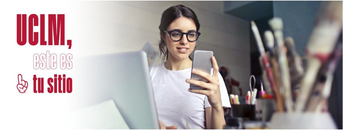 Mujer joven sentada delante de un ordenador portátil sonríe mientras mira un teléfono móvil 